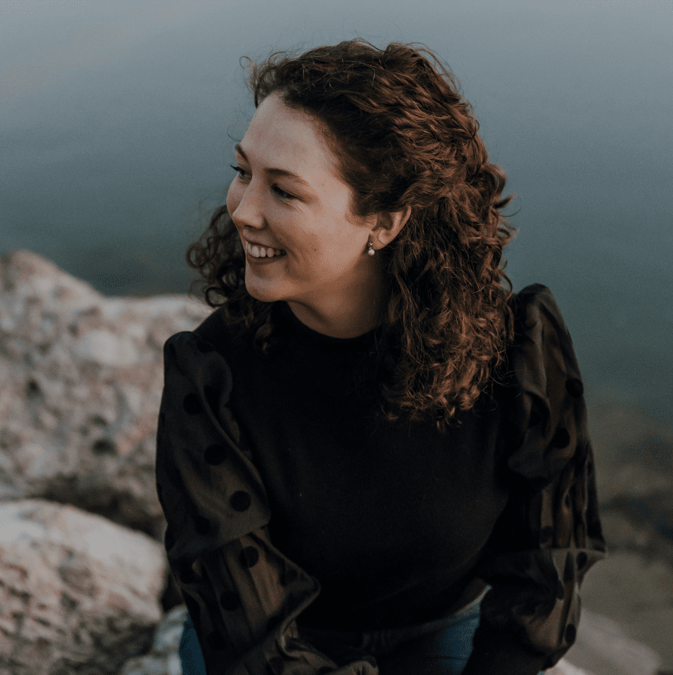 Eine junge Frau, Veronika Hilger, mit lockigem Haar sitzt lächelnd auf hellen Felsen am Wasser. Sie trägt ein schwarzes Oberteil mit transparenten Ärmeln und blickt seitlich, während das weiche Licht eine ruhige und warme Stimmung erzeugt. Das Bild vermittelt Natürlichkeit, Vertrauen und Geborgenheit – passend zum Thema Erstgespräch für persönliche Wochenbettbetreuung | Unterstützung im Wochenbett für Mamas | Begleitung nach der Geburt