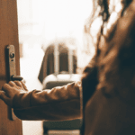 Eine Person öffnet eine Haustür, im Hintergrund steht ein gepackter Koffer in einem hellen Zimmer. Symbolbild für angekündigten oder unangekündigten Besuch im Wochenbett.