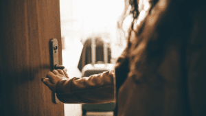 Eine Person öffnet eine Haustür, im Hintergrund steht ein gepackter Koffer in einem hellen Zimmer. Symbolbild für angekündigten oder unangekündigten Besuch im Wochenbett.