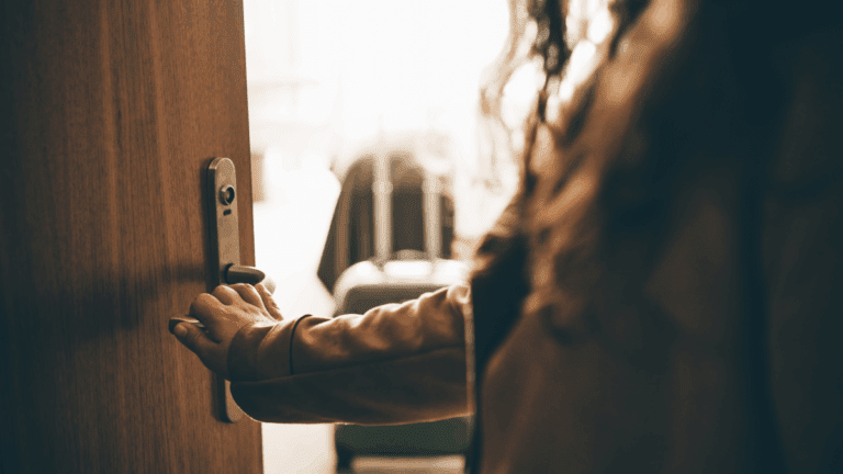 Eine Person öffnet eine Haustür, im Hintergrund steht ein gepackter Koffer in einem hellen Zimmer. Symbolbild für angekündigten oder unangekündigten Besuch im Wochenbett.